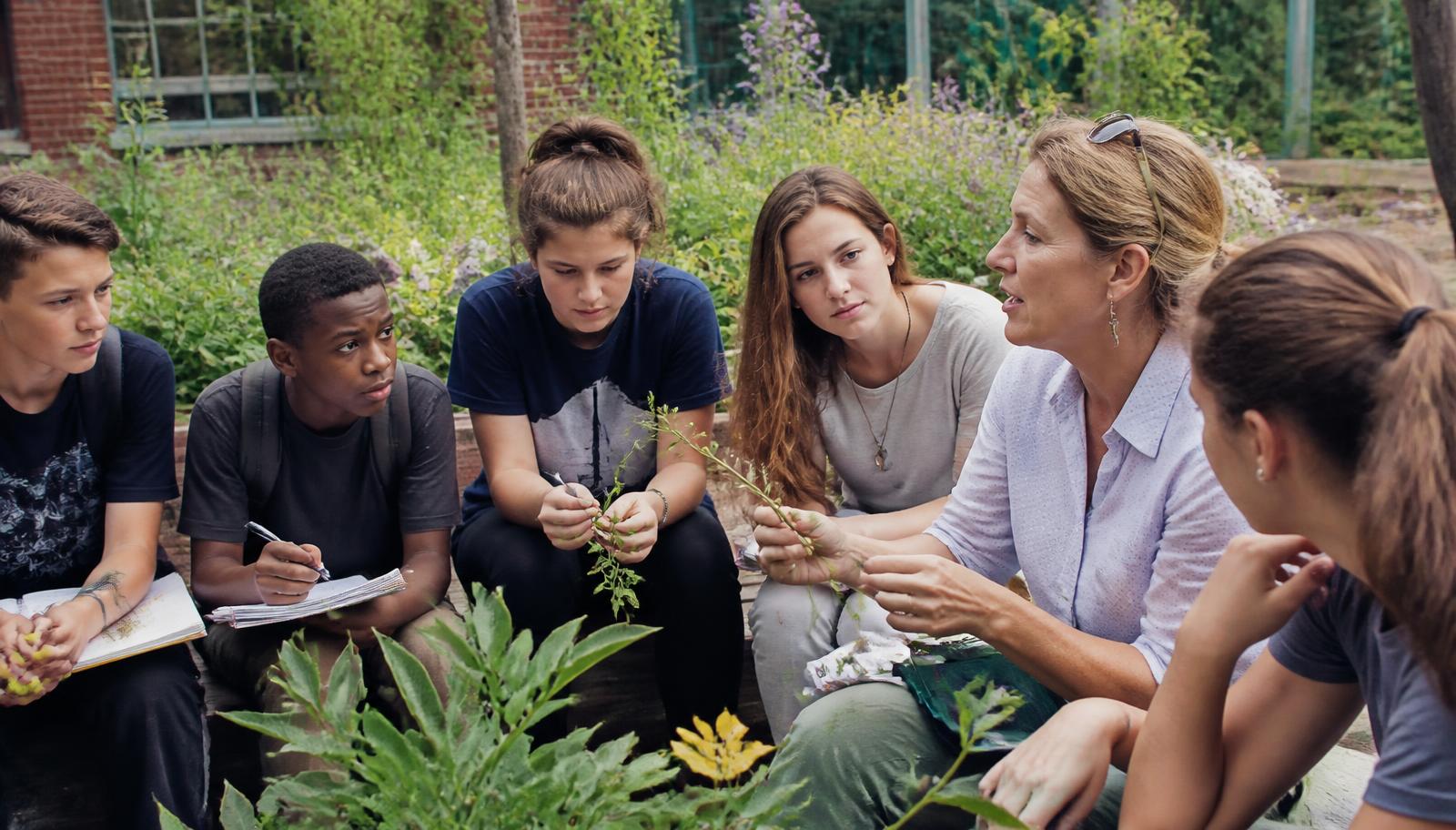 Young people and an educator studying plant samples in an outdoor classroom beside a garden